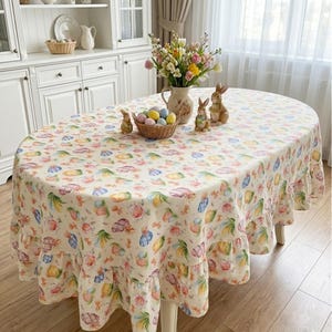 May include: Oval Easter tablecloth with a cream background and a repeating pattern of colorful Easter eggs, flowers, and bows. The tablecloth has a ruffled edge and is set on a white table. Decorative Easter bunnies and a basket of eggs are on the table.