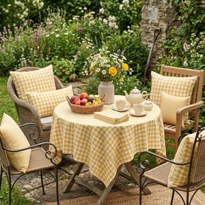 May include: A charming outdoor dining scene with a round table covered in a yellow and white gingham tablecloth. Wicker and wooden chairs with matching gingham cushions surround the table, set with a teapot, cups, and a basket of fruit. A vase of flowers adds a touch of color.