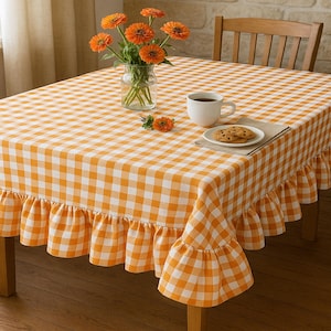 Puede incluir: Un mantel a cuadros naranja y blanco con un borde con volantes cubre una mesa de madera. Un jarrón con flores naranjas, una taza de café y un plato de galletas están sobre la mesa. El mantel tiene un patrón de cuadros.