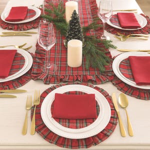 Pode incluir: Um conjunto de mesa com um caminho de mesa xadrez vermelho e verde e individuais com folhos a condizer. Cada prato branco &eacute; coberto por um guardanapo vermelho liso e talheres dourados. Duas velas brancas e um pequeno pinheiro.
