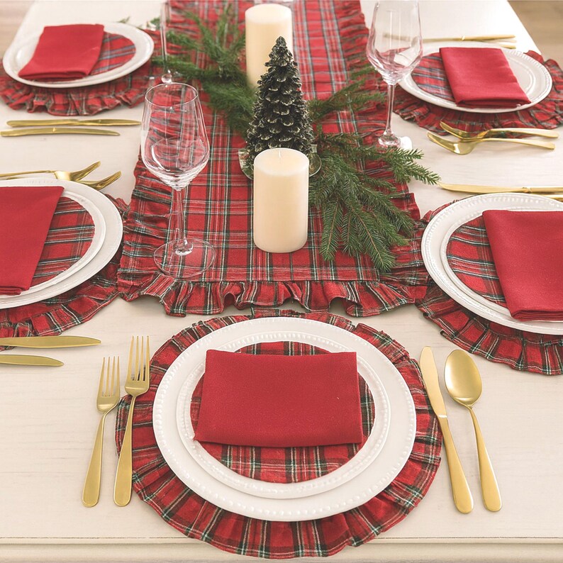 Pode incluir: Um conjunto de mesa com um caminho de mesa xadrez vermelho e verde e individuais com folhos a condizer. Cada prato branco &eacute; coberto por um guardanapo vermelho liso e talheres dourados. Duas velas brancas e um pequeno pinheiro.