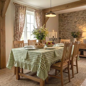 May include: A rectangular dining table covered with a light green and white checkered tablecloth featuring a ruffled edge. The table is set with white plates, a woven basket filled with bread, a bowl of fruit, and a black metal candelabra.