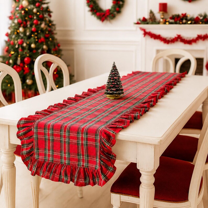 Pode incluir: Um caminho de mesa retangular com um padr&atilde;o xadrez em vermelho, verde e branco, com bordas franzidas nas laterais. O caminho de tecido est&aacute; estendido sobre uma mesa de jantar branca, com uma pequena &aacute;rvore de Natal artificial decorativa.