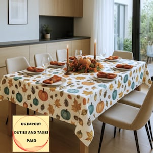 Puede incluir: Una mesa de comedor preparada para una ocasión festiva, con un mantel blanco adornado con calabazas coloridas y hojas de otoño. La mesa está puesta con platos, servilletas y copas de vino, con un centro de mesa de calabaza y velas encendidas.