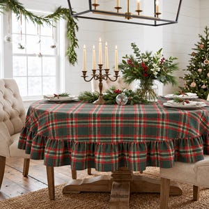 May include: A round tablecloth featuring a dark green, red, and white plaid pattern with a ruffled edge. The tablecloth is draped over a wooden table set with a five-arm metal candelabra, white taper candles, and a glass vase of greenery.