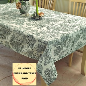 Op de afbeelding: Een rechthoekig tafelkleed met een gedetailleerd groen en wit bloemenpatroon. De stof drapeert over een houten tafel en toont het ingewikkelde ontwerp. Het tafelkleed heeft een klassieke toile de Jouy-stijl en voegt een vleugje elegantie toe.