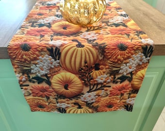 May include: A fall-themed table runner with a pumpkin and floral pattern. The runner features orange, yellow, and white flowers and pumpkins. A gold-colored glass pumpkin sits on top. Perfect for autumn decor.