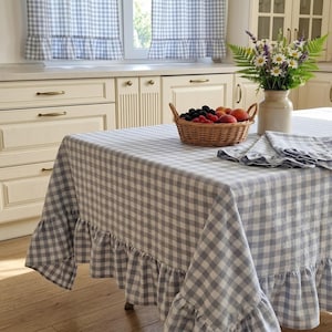 Puede incluir: Un mantel a cuadros grises y blancos con un borde con volantes cubre una mesa. Una cesta de mimbre llena de fruta y un jarrón con flores están sobre la mesa. Cortinas a cuadros a juego en el fondo.