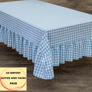 Puede incluir: Un mantel a cuadros vichy azul claro y blanco con un borde con volantes. El mantel rectangular cubre una mesa. La tela tiene un patrón de cuadros clásico, creando una estética vintage.