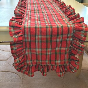Pode incluir: Um caminho de mesa retangular com um padr&atilde;o xadrez em vermelho, verde e dourado. O caminho de mesa &eacute; rematado com um folho franzido ao longo de todo o per&iacute;metro, criando uma borda decorativa que emoldura o design central de tecido xadrez.