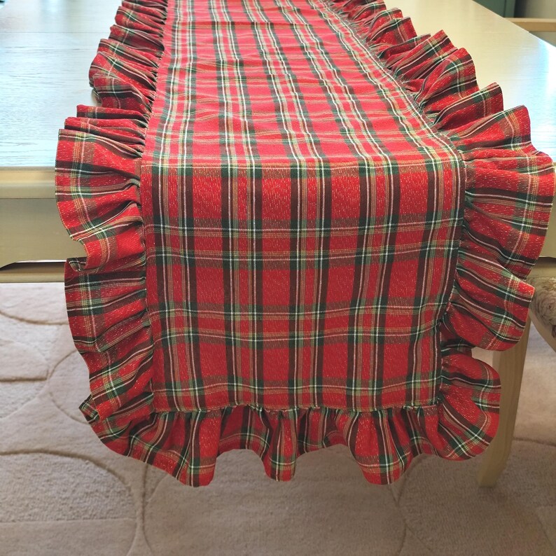 Pode incluir: Um caminho de mesa retangular com um padr&atilde;o xadrez em vermelho, verde e dourado. O caminho de mesa &eacute; rematado com um folho franzido ao longo de todo o per&iacute;metro, criando uma borda decorativa que emoldura o design central de tecido xadrez.