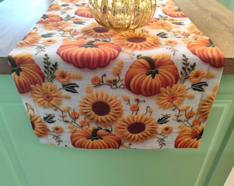 May include: A fall-themed table runner with a repeating pattern of orange pumpkins and yellow sunflowers on a cream background. A gold-colored glass pumpkin sits on top of the runner. The runner is displayed on a wooden surface.