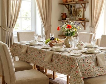 Puede incluir: Una mesa de comedor con un mantel de estampado floral, sillas tapizadas en beige y un jarrón de cerámica con flores silvestres. La mesa incluye platos blancos, cristalería, una tetera y una cesta de pan en una habitación luminosa.