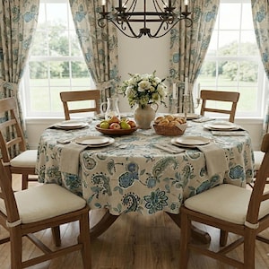 May include: A round dining table set with a beige tablecloth featuring a blue and green floral paisley pattern. The table includes plates, napkins, a bowl of apples, a basket of bread, and a vase of white flowers, surrounded by wooden chairs.
