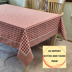May include: A red and white checkered tablecloth covers a wooden table. The tablecloth has a wide border of smaller red and white checks. The table is set in a room with wooden chairs and a neutral-colored carpet.