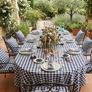 May include: An outdoor dining table set for a meal. The table is covered with a blue and white checkered tablecloth and set with plates, silverware, and glasses. Chairs with matching checkered cushions surround the table. A floral centerpiece and candles add to the ambiance.