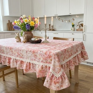 May include: A rectangular tablecloth with a soft pink and white floral pattern, featuring a ruffled skirt edge trimmed with white lace. The table is set with a brown ceramic vase of colorful spring flowers, two ceramic bunnies, and a candle holder.