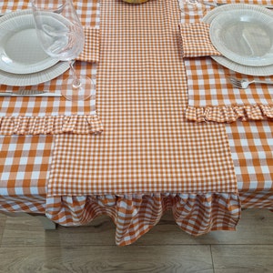 Op de afbeelding: Oranje en wit geruite tafelkleed met gekrulde randen. Twee couverts met witte borden en een glas staan op het tafelkleed.