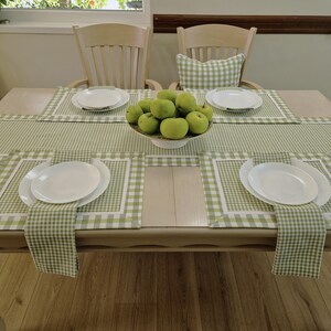 May include: A green and white gingham tablecloth with four place settings. Each place setting includes a white plate, a green and white gingham napkin, and a green and white gingham placemat. A bowl of green apples sits in the centre of the table.