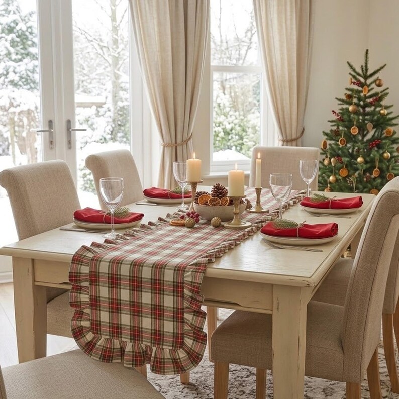 Pode incluir: Um caminho de mesa retangular com um padr&atilde;o xadrez em vermelho, verde e creme com bordas franzidas. O caminho est&aacute; colocado sobre uma mesa de jantar de madeira clara com pratos brancos, guardanapos de tecido vermelhos e velas.