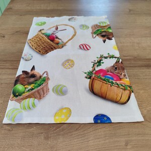 Op de afbeelding: Een witte keukendoek met een kleurrijk paasmotief. De handdoek toont konijnen in manden met kleurrijke eieren. De achtergrond is wit met een verspreid patroon van kleurrijke eieren.