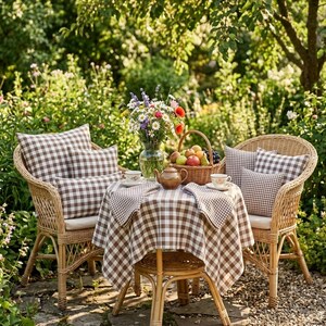 May include: Two wicker chairs with brown and white checkered pillows and a small table set for tea in a garden. A checkered tablecloth covers the table, with a teapot, cups, and a basket of fruit. A vase of flowers adds color.