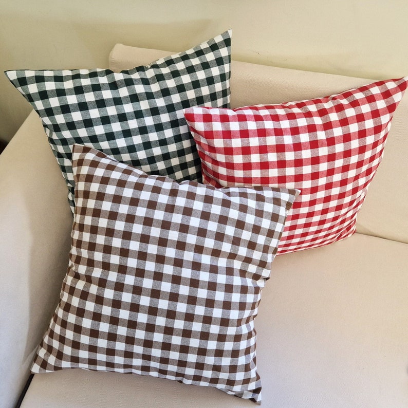 May include: Three square throw pillows in a variety of colors, arranged on a beige sofa. The pillows feature a classic gingham pattern in green and white, red and white, and brown and white. The pillows are soft and inviting.