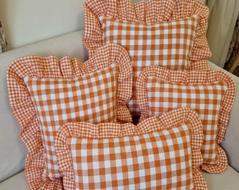 Könnte beinhalten: Eine Sammlung von drei quadratischen und rechteckigen Dekokissen mit einem orange-weißen Vichy-Karo-Muster. Jedes Kissen ist mit einem gerüschten Stoffrand versehen, der die Kanten einfasst und für ein dekoratives und strukturiertes Aussehen sorgt.
