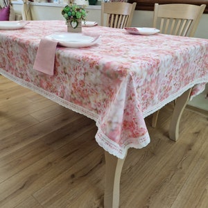 May include: A pink floral tablecloth with a white lace trim. The tablecloth is laid on a wooden table with two chairs.