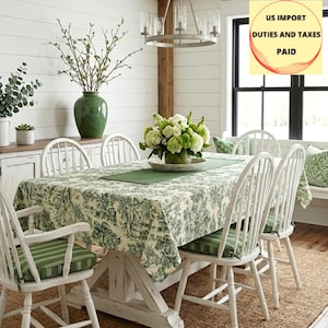 Op de afbeelding: Een eethoek met een witte boerderijtafel en bijpassende stoelen. De tafel is gedekt met een groen-wit bloemenkleed, een groene loper en een bloemstuk. De stoelen hebben groen gestreepte kussens.