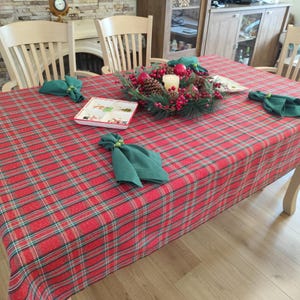 May include: Red and green plaid tablecloth with a Christmas centerpiece of a green wreath with red berries, pine cones, and a white candle. Four green napkins are folded on the table.