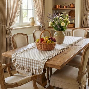 May include: A rustic dining room scene featuring a wooden table set with a checkered table runner and a basket of fruit. The room is decorated with beige curtains and a vase of flowers, creating a warm, inviting atmosphere.