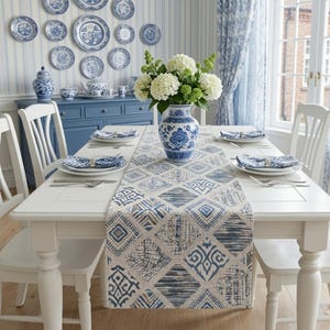Op de afbeelding: Een eettafel gedekt met blauw en wit porselein en een patroon tafelloper. Een vaas met witte bloemen staat in het midden. De kamer is ingericht met blauw en wit gordijnen en wandborden, wat een samenhangende esthetiek creëert.