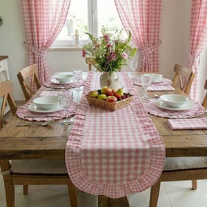 Pode incluir: Uma mesa de jantar rústica de madeira, preparada para uma refeição. A mesa é adornada com um caminho de mesa e individuais de xadrez rosa e branco. Taças brancas, copos e um cesto de frutas completam a composição. Cortinas xadrez rosa emolduram a janela.