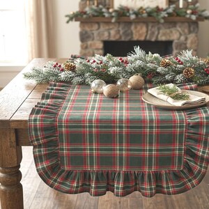 Pode incluir: Um caminho de mesa retangular com padr&atilde;o xadrez em verde escuro, vermelho e creme com uma borda franzida. O caminho est&aacute; estendido sobre uma mesa de madeira, decorado com uma guirlanda de pinheiro geada, pinhas e bagas vermelhas.