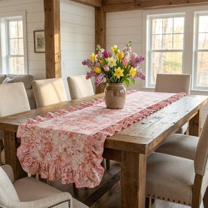 May include: A rustic wooden dining table is set with a pink floral patterned table runner with ruffled edges. A ceramic vase holds a bouquet of colorful flowers. Beige upholstered chairs surround the table, creating a warm, inviting dining space.
