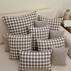 Peut inclure: Plusieurs coussins à carreaux marron et blanc de différentes tailles, disposés sur un canapé beige. Les coussins présentent un motif vichy classique, créant une ambiance chaleureuse et accueillante. L'arrangement comprend des formes carrées et rectangulaires.
