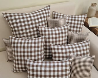Könnte beinhalten: Eine Sammlung braun-weiß karierter Zierkissen in verschiedenen Größen, die auf einem beigen Sofa angeordnet sind. Die Kissen haben ein klassisches Vichy-Muster und schaffen eine gemütliche und einladende Atmosphäre. Die Anordnung umfasst quadratische und rechteckige Formen.