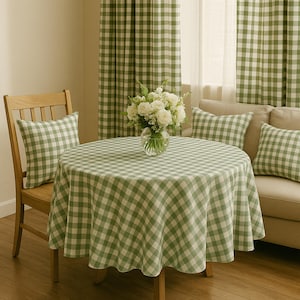 May include: A round table covered with a green and white gingham tablecloth, featuring a glass vase filled with white roses. The setting includes a wooden chair and a sofa, both adorned with matching green and white gingham throw pillows.
