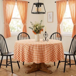 May include: A dining room with a round wooden table covered in an orange and white gingham tablecloth. Black chairs surround the table. Orange gingham curtains frame the windows. A vase of flowers and a candle holder are on the table.