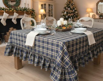 May include: A square tablecloth featuring a dark blue, green, and yellow plaid pattern with a ruffled edge. The fabric is draped over a wooden table set with white bowls, clear glasses, and beige napkins, creating a coordinated dining arrangement.