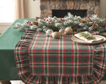 Pode incluir: Um caminho de mesa retangular com um padrão xadrez em verde escuro, vermelho e dourado com uma borda franzida. O caminho é colocado sobre uma toalha de mesa verde escura lisa e decorado com uma guirlanda de ramos de pinheiro, bagas vermelhas e pinhas.