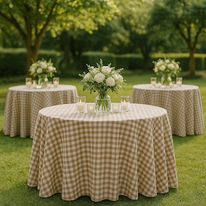 May include: Three round tables with tan and white checkered tablecloths are set up on a green lawn. Each table has a vase of white roses and several lit candles. The scene is outdoors with trees in the background.