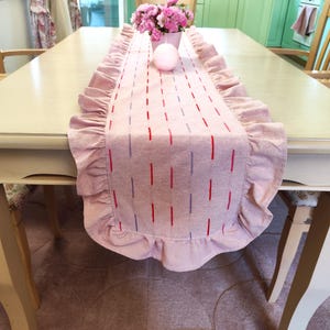 Può includere: Una tovaglia rosa con bordi a balze e linee verticali ricamate rosse e viola. La tovaglia è posta su un tavolo di legno chiaro. Un vaso di fiori rosa e una candela rosa sono sul tavolo.