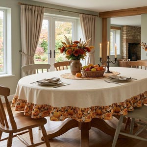 May include: An oval dining table covered with a beige tablecloth featuring a ruffled edge with a pumpkin and autumn leaf print. The table holds a ceramic vase with fall flowers, a woven basket of fruit, and a black metal candelabra.