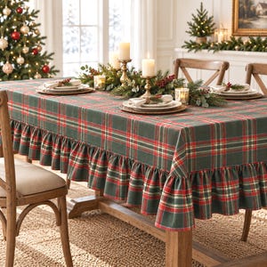 May include: A rectangular tablecloth featuring a dark green, red, and white plaid pattern with a gathered ruffle trim along the edges. The fabric covers a wooden table set with white plates, napkins, and a centerpiece of greenery and candles.
