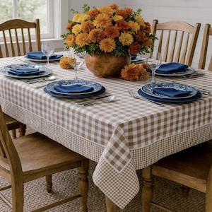 Pode incluir: Uma mesa de jantar montada para uma refeição, com uma toalha de mesa xadrez marrom e branca. A mesa está montada com pratos azuis, guardanapos, talheres e taças de vinho. Um grande arranjo floral de flores laranja e amarelas está no centro.