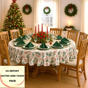 Puede incluir: Una mesa de comedor festiva para una comida navideña. La mesa redonda está cubierta con un mantel blanco con un estampado botánico rojo y verde. Se colocan servilletas y platos verdes para cada comensal, con velas rojas y un centro de mesa floral.