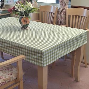 May include: A green and white gingham tablecloth on a wooden table. A vase of flowers sits on the table. Two wooden chairs are in the background, creating a homely dining setting. The table is approximately 150 cm long.