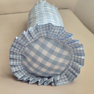 Könnte beinhalten: Ein blau-weiß kariertes Nackenrolle-Kissen mit gerüschtem Rand. Das Kissen ist zylindrisch mit einem abgerundeten Ende und einer Rüschenverkleidung. Der Stoff hat ein klassisches Karomuster.